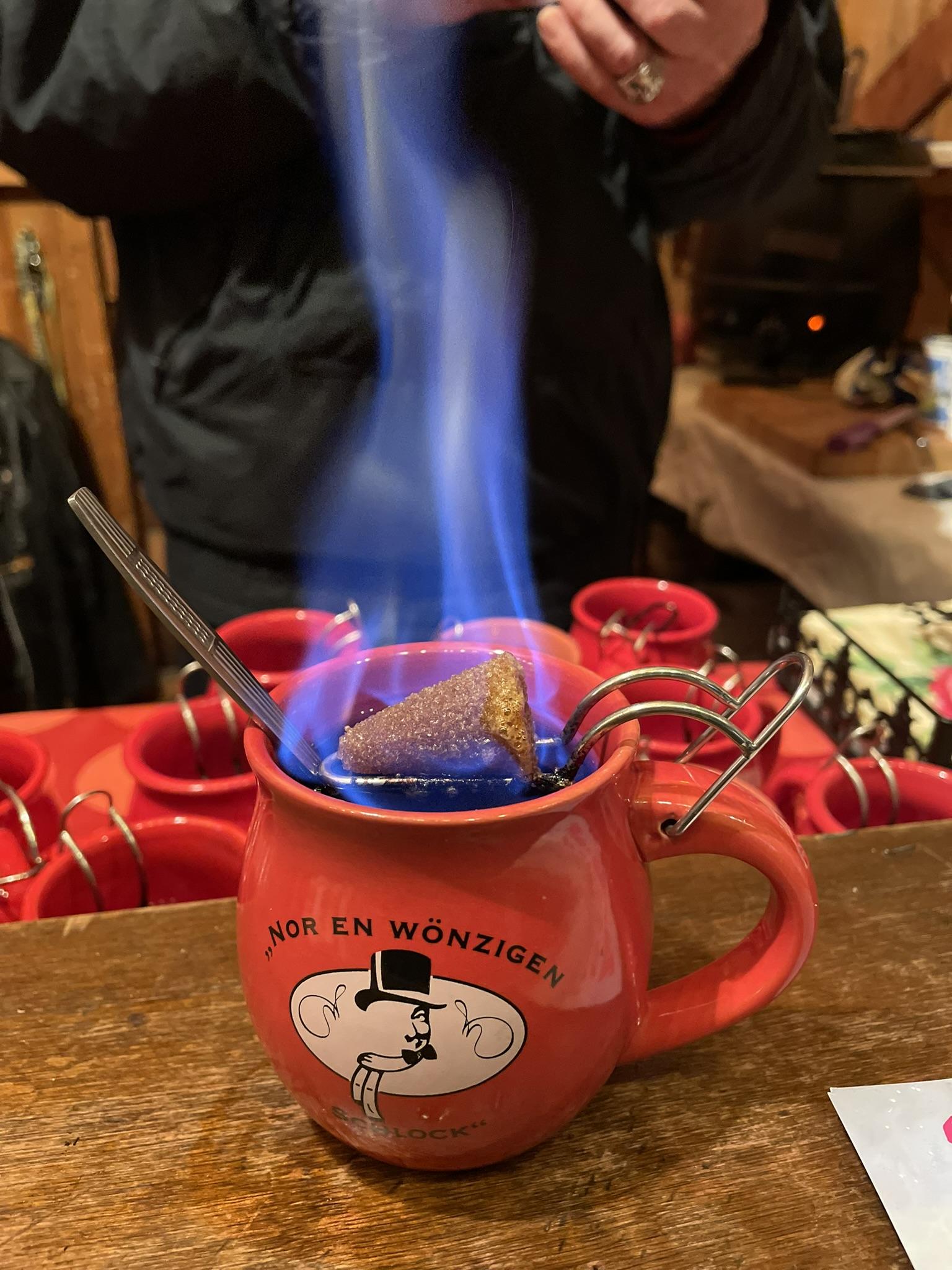 Ein roter Becher mit gewürztem, warmem Rotwein. Darauf eine Feuerzange mit einem in Rum getränkten, kegelförmigen Zuckerstück. Das brennt mit einer hohen blauen Flamme und tropft in die Flüssigkeit. 