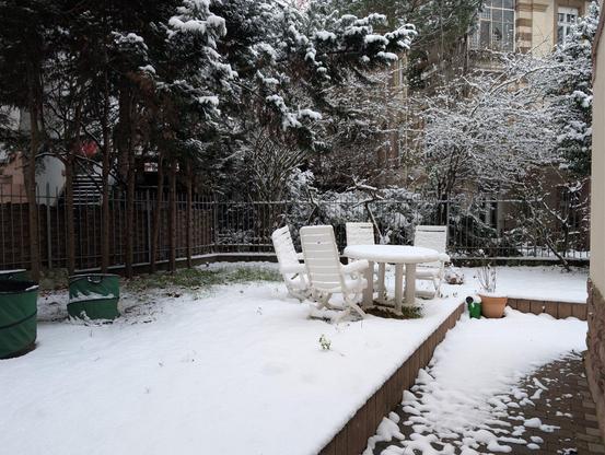 Ein von Schnee bedeckter Garten. Im Zentrum ein weißer Tisch mit vier weißen Gartenstühlen. Drumherum verschiedene Sträucher und Nadelbäume.