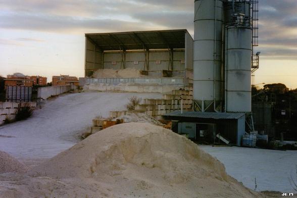 Farbfoto, aufgenommen gegen Sonnenuntergang, das eine bautechnische Anlage mit Sand im Vordergrund und zwei Silos rechts weiter hinten zeigt. Links neben den Silos ein vorne offenes Lagergebäude aus Blech.