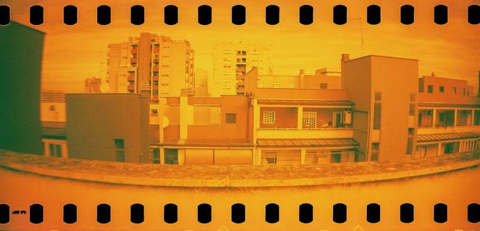 Farbfoto im Rot-Orange-Farbspektrum, das eine stark verzerrte panoramische Ansicht von Wohngebäuden in Rom zeigt. Aufgenommen mit einer Sprocket Rocket-Kamera, bei der auch die Führungslöcher des Films Teil des Bildes sind.
