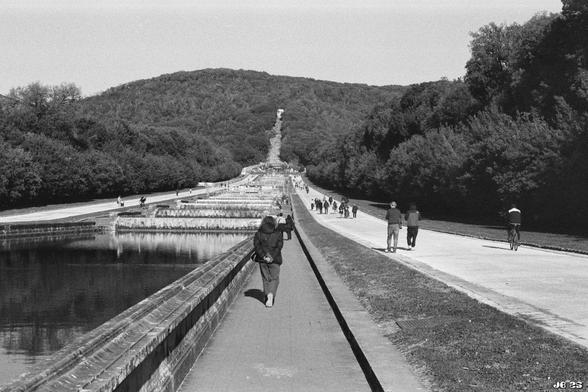 Schwarzweißfoto vom Park der Reggia di Caserta in Italien. Zu sehen ist eine lange Promenade mit etlichen Besuchern im Barockgarten. Am Horizont der Bergpark, den es zu erreichen gilt. Leica R4 mit 50 mm Summilux auf Potsdam-Schwarzweißfilm.