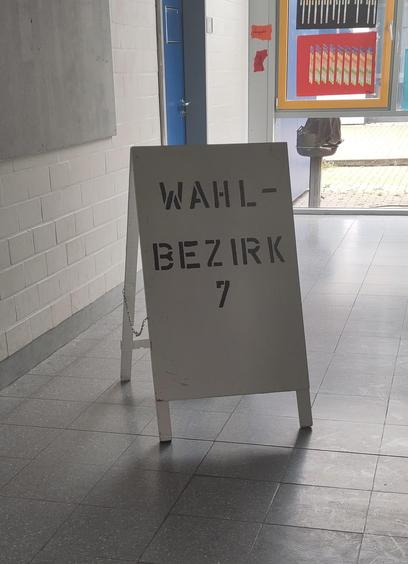 Ein grau gefliester Gang in einer Schule, in der Mitte des Bildes eine weiße Klapptafel mit der Aufschrift "WAHLBEZIRK 7"