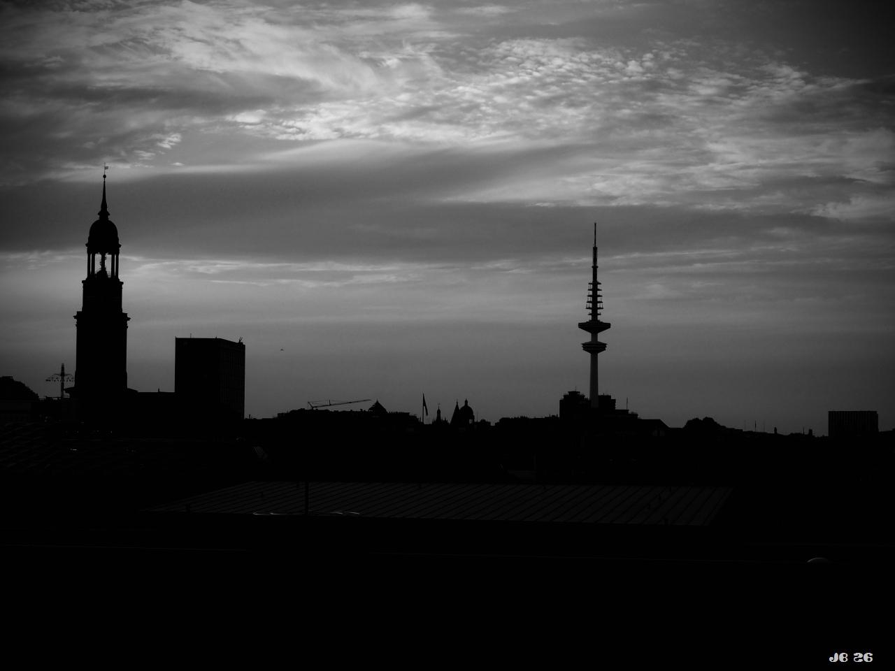 Dunkles Schwarzweißfoto mit einem Blick über Hamburg von der Elbphilharmonie aus. Himmel mit bewegtem Wolkenmuster, darunter scherenschnittartig hohe Gebäude, darunter der Turm der Michaeliskirche und der Fernsehturm, die aus der Stadt herausragen.