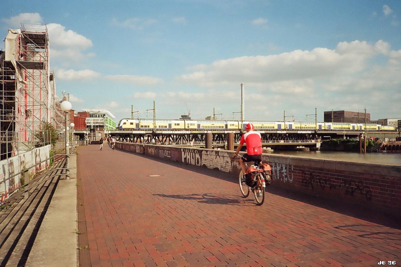 Farbfoto. Ein Fahrradfahrer fährt in Hamburg auf einem gepflasterten Weg an einem Kanal entlang, den ein Zug auf einer Brücke überquert. Am hellblauen Himmel ein paar Wolken. Olympus XA auf Kodakcolor 200-Film.