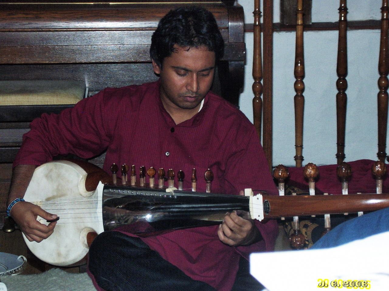 Ein indischer Musiker sitzt auf einer weißen Bank und spielt seine Sarod. Im Hintergrund links ein Klavier, rechts ein Geländer aus gedrechselten Stäben.