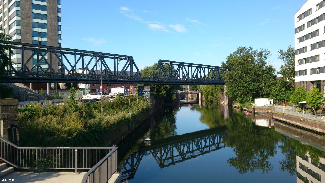 Farbfoto mit Blick auf einen Kanal in Hamburg mit einer querenden Eisenbahnbrücke, die sich im Wasser spiegelt. Links und rechts Gebäude in Vegetation.
