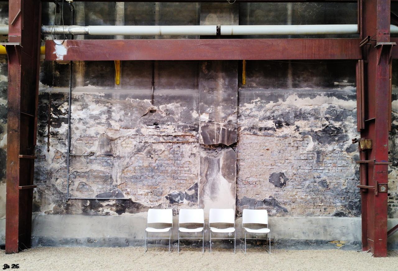Farbfoto, aufgenommen in einer ehemaligen Werkshalle in Büdelsdorf, die jetzt als Raum für Kunstausstellungen dient. Vor einer lädierten Backsteinmauer stehen zentriert vier weiße Stühle. Umrahmung oben, links und rechts durch ein rostfarbenes Metallgerüst.