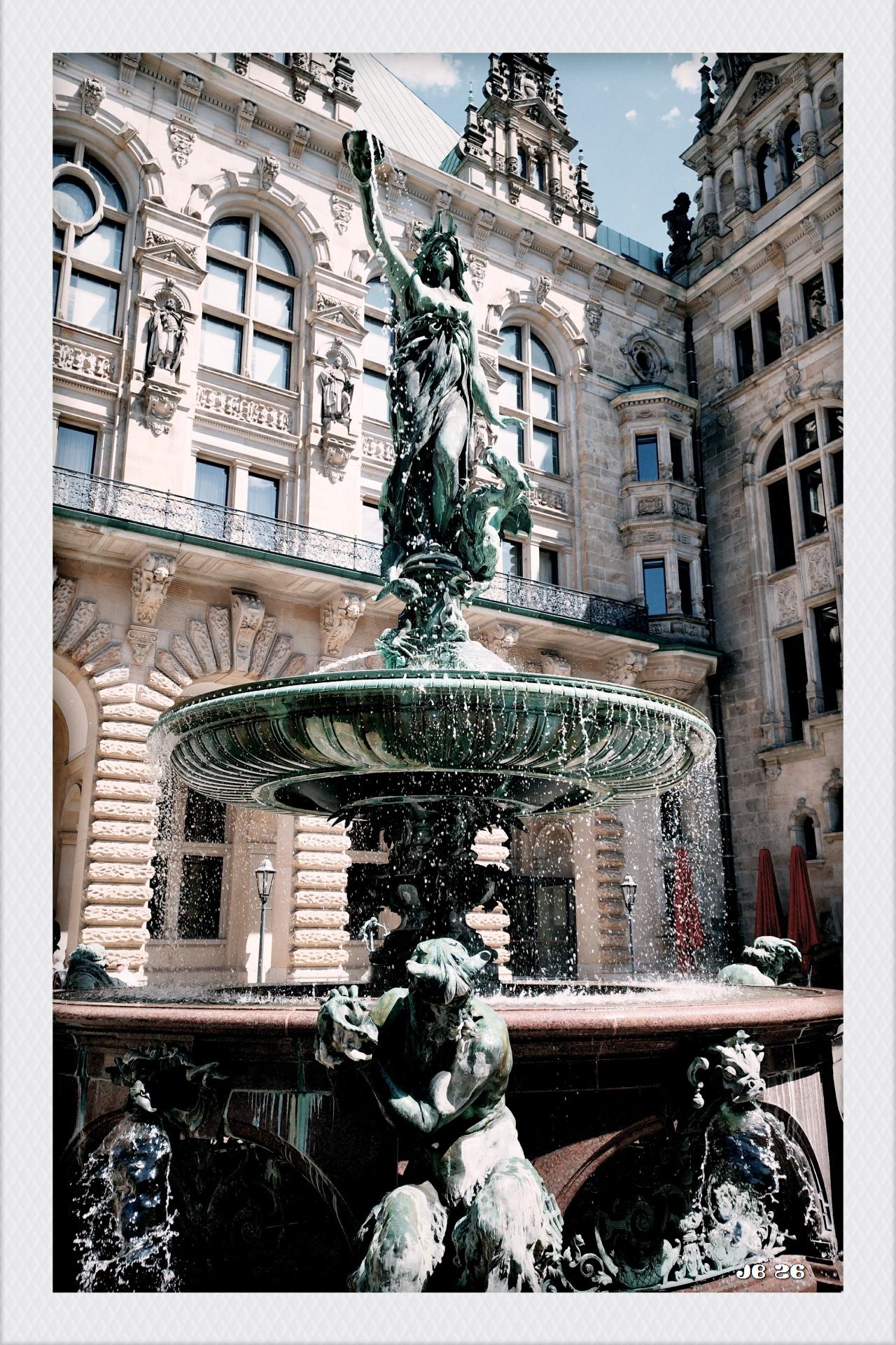 Farbfoto in etwas kühlen Farben mit Blick auf den Hygieia-Brunnen im Innenhof des Hamburger Rathauses, das im Hintergrund zu sehen ist. Oben auf dem Brunnen die Statue der Göttin Hygieia, die auf einer breiten Schale steht, von der das Wasser in den eigentlichen Brunnen überläuft. Unten vor dem Brunnen eine Bronzefigur.