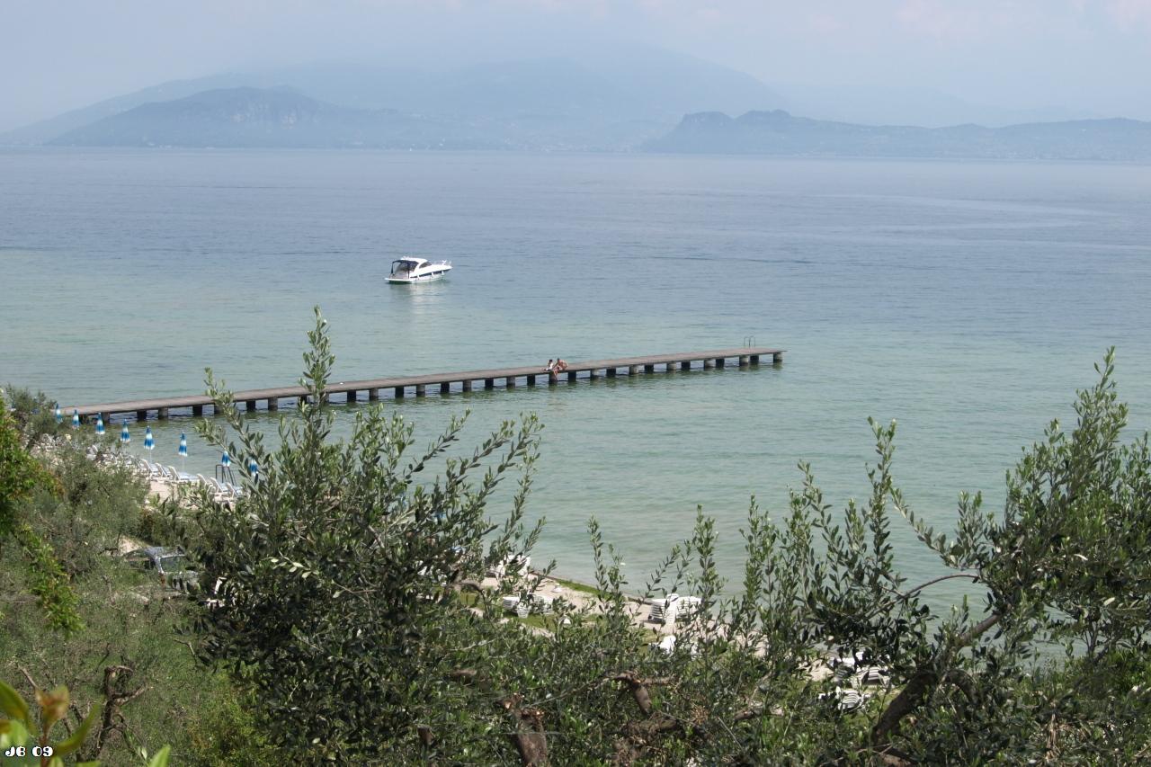 Blick auf das Wasser des Gardasees von oberhalb über Gebüsch. Im Wasser ein Steg, weiter entfernt ein Motorboot, am Horizont verschwommen Berge und Himmel.