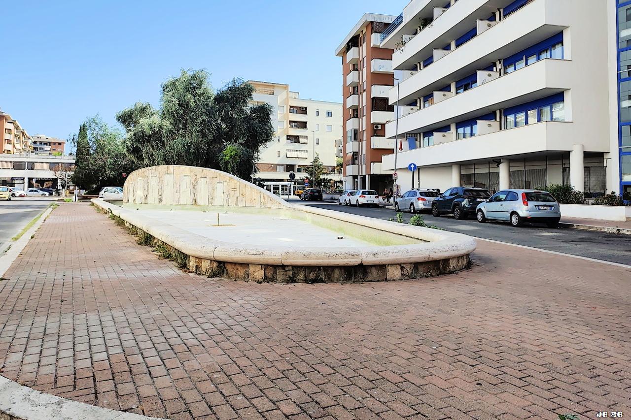 Farbfoto mit Blick auf einen länglich-ovalen Brunnen in Bildmitte, der wegen der lang anhaltenden Hitze kein Wasser enthält. Wurde vermutlich wegen Wasserknappheit abgeschaltet. Um den Brunnen rötliches Plaster, rechts und weiter entfernt große Wohngebäude. Hinter dem Brunnen grüne Bäume.