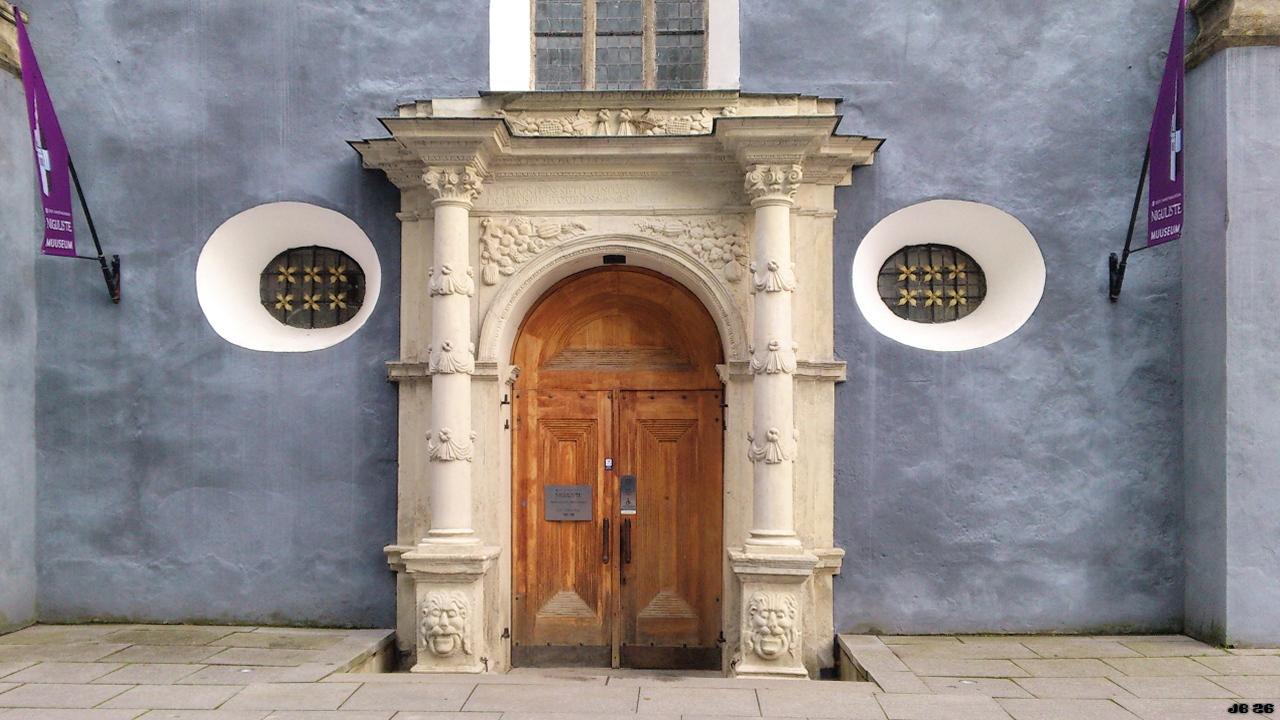 Farbfoto mit Blick auf den Eingang einer Barockkirche in Talinn, Estland. In Bildmitte die geschnitzte Tür aus hellem Holz, umrahmt von einem Portal aus hellem Stein. Darüber ein Fenster, links und rechts zwei weiß vertiefte runde Fenster mit Verzierung. Weiter rechts und links lila Flaggen.
