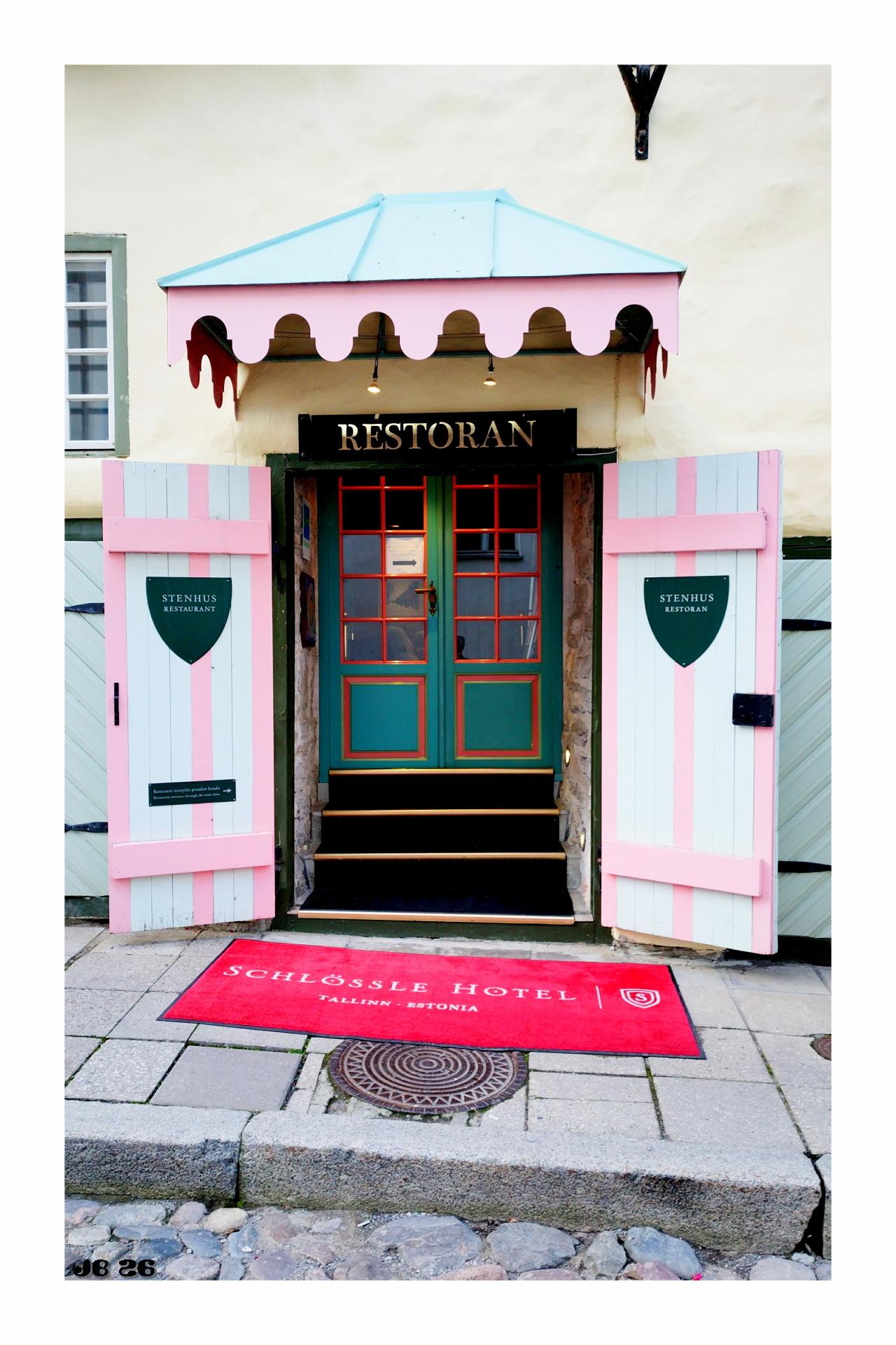 Foto im Hochformat, das den Eingang eines Hotel-Restaurants in Talinn, Estland, zeigt, der hauptsächlich in Pastellfarben (rosa und hellblau) gehalten ist. Zwei Flügeltüren und ein Vordach zur Dekoration. Drei Stufen führen zur Eingangstür hoch, deren Farben kräftiger sind.