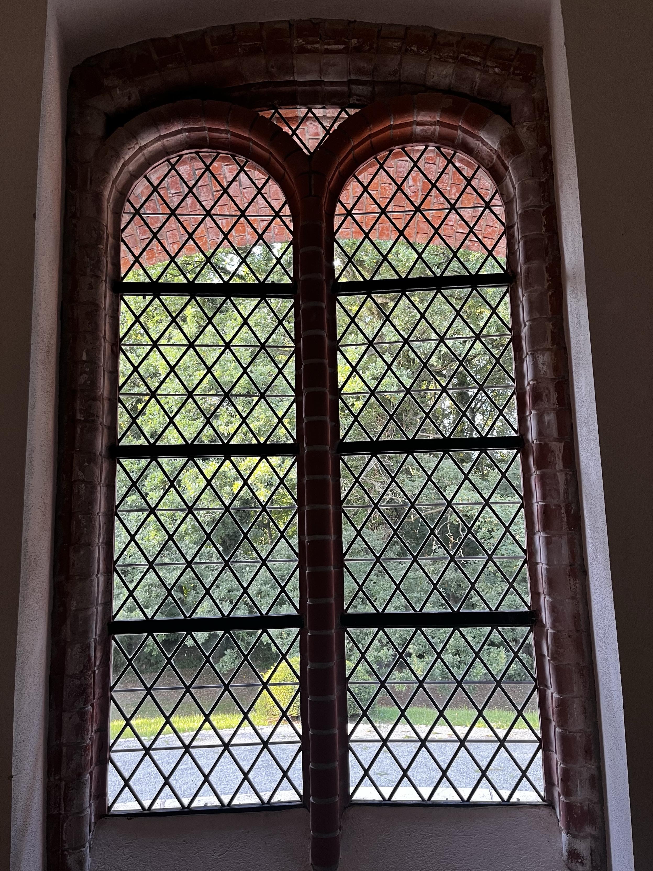 A large, arched window with brick framing and diamond-patterned glass panes. The view shows greenery and a paved area outside.