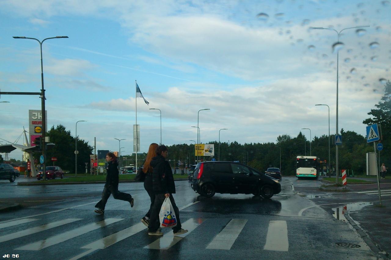 Vor einem Zebrastreifen aus dem stehenden Auto aufgenommenes Farbfoto an einem Regentag, das drei Personen zeigt, die die Straße überqueren. Die Frau links läuft schnell. Weiter entfernt ein querendes Auto, am Horizont ein Bus, eine Tankstelle, Wald, der wolkenbehangene blaue Himmel und Regentropfen auf der Windschutzscheibe.