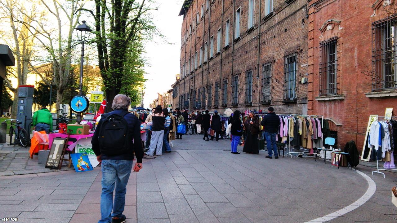 Farbfoto mit Blick auf einen Straßenmarkt in Piacenza, Emilia Romagna, Italien, mit verschiedenen Verkaufsständen vor dem Backsteingebäude rechts und einer Parkanlage links. Der Markt zieht einige Besucher an.