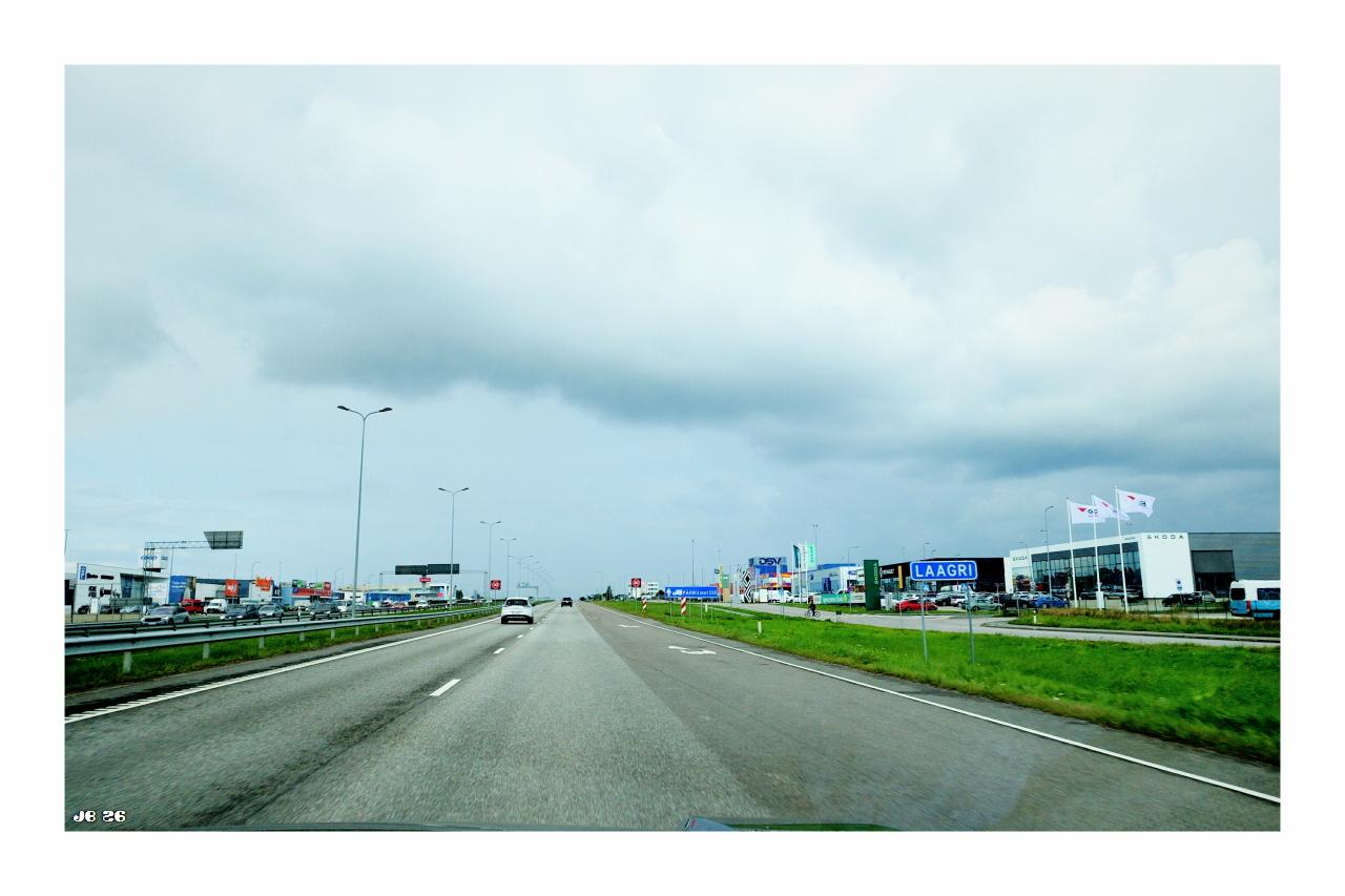 Weitwinkel-Farbfoto, aufgenommen durch die Windschutzscheibe eines fahrenden Autos in Estland an einem bewölkten Tag. Links und rechts von einer breiten Straße etliche Gebäude und Masten. Blaugrauer Himmel mit weißlichen Wolken.