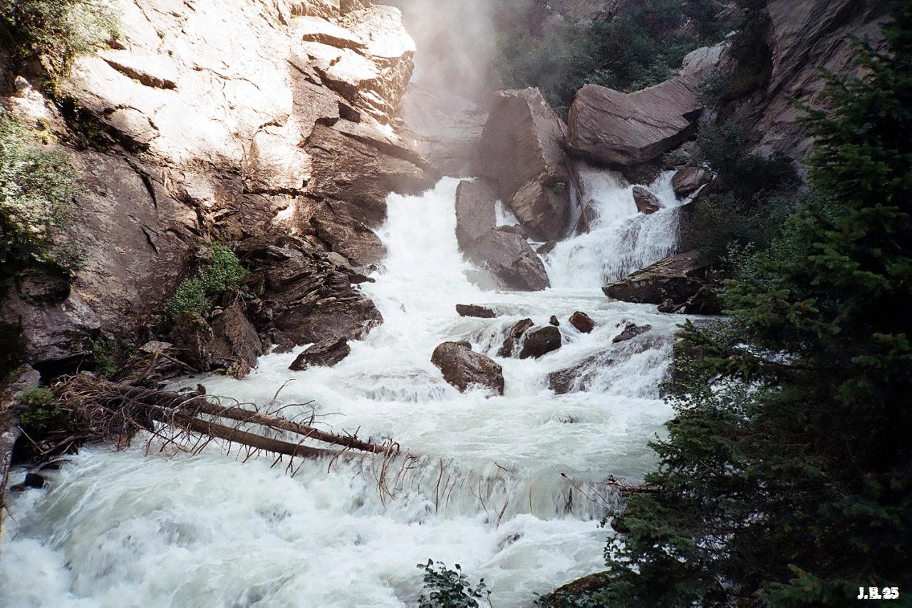 Farbfoto eines reißenden Alpenflusses im Aostatal, der hier wasserfallartig zwischen Felsbrocken und umgestürzten Bäumen abfällt. Links ein großer Felsen, rechts ein Nadelbaum. Olympus XA auf Kodak Gold.