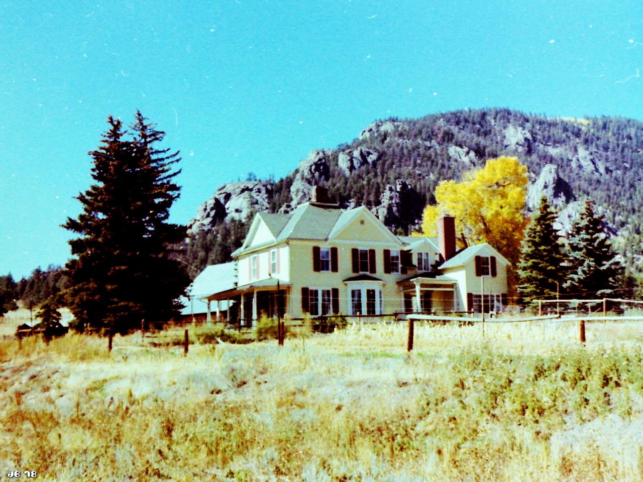 Verblichenes Farbfoto, aufgenommen in den Rocky Mountains in Colorado. In Bildmitte ein großes hellgelbes Haus, links und rechts davon Bäume, darunter auch eine Pappel (Aspen) im gelben Herbstkleid. Im Hintergrund ein bewaldeter Berg und blauer Himmel.