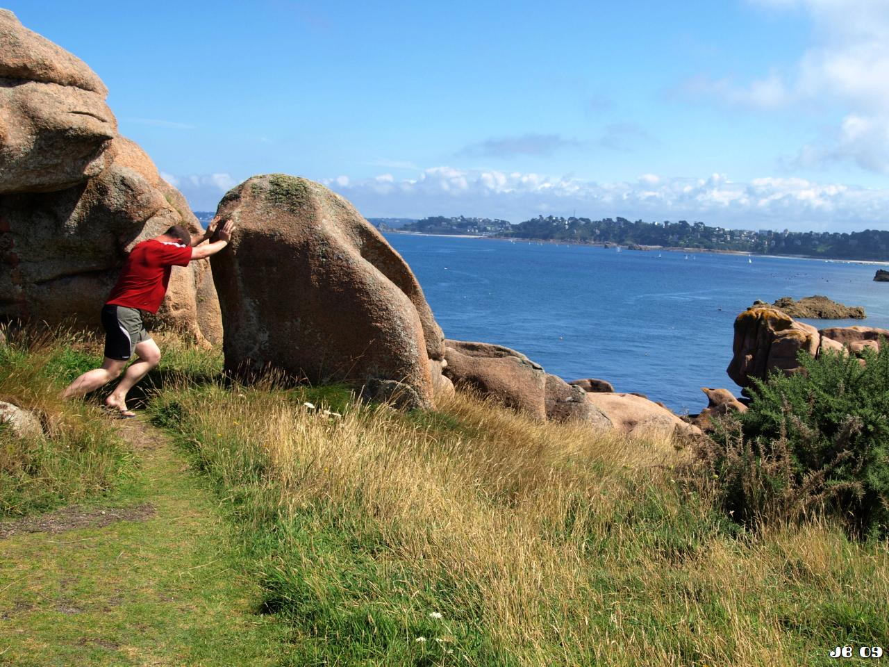 Farbfoto, aufgenommen an der Küste in der Bretagne. Ein Mann tut so, als ob er einen großen Felsen bewegen könnte.