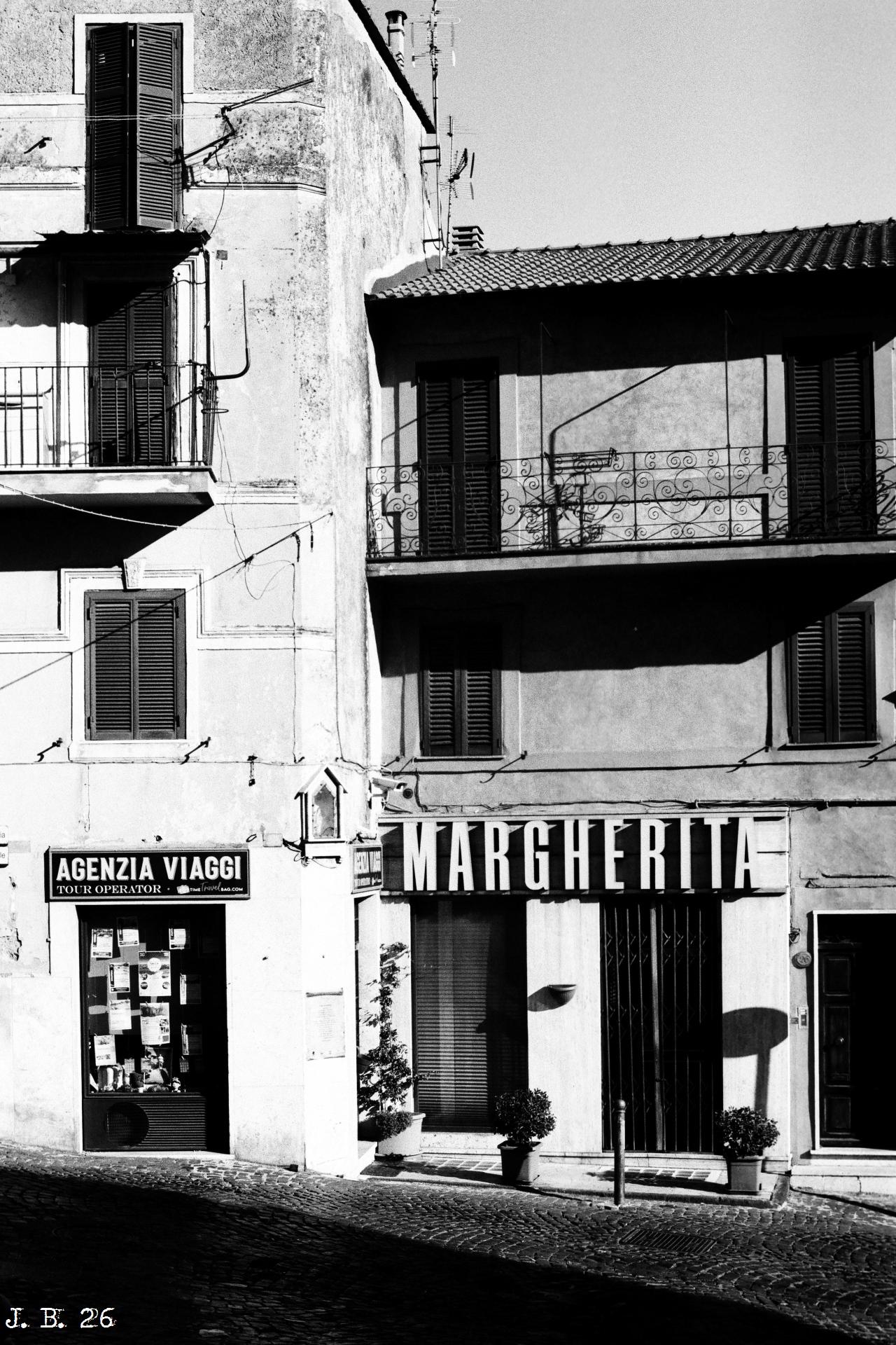 Schwarzweißfoto der Fassaden von zwei aneinandergebauten Häusern in Monte Compatri, Lazio, Italien. Im linken, vierstöckigen Haus ist unten ein Reisebüro untergebracht, im rechten Haus ein Laden namens "Margherita". Hoher Kontrast und starker Schattenschlag. Leica R4 mit 50 mm Summilux auf Lomo Earl Grey-Film.