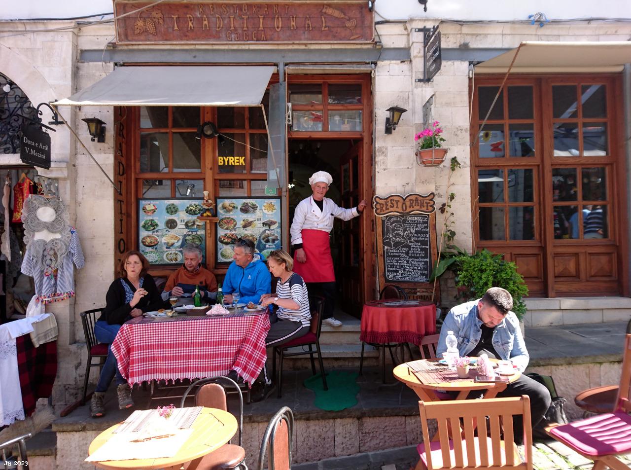 Farbfoto mit Blick auf ein Restaurant irgendwo in Albanien. Im Vordergrund Tische, zwei davon mit Gästen, in Bildmitte vor der Eingangstür der Besitzer mit roter Schürze, weißem Hemd und weißer Kochmütze. Links und rechts Fenster des Lokals in quadratischen Holzrahmen, rechts vom Koch das Tagesmenü.