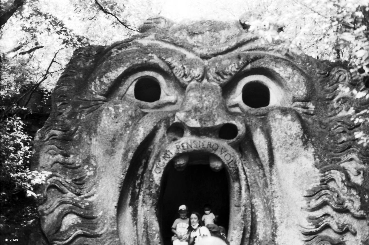 Schwarzweißfoto, das den Kopf eines Monsters aus Stein mit weit aufgerissenem Mund zeigt. Im Mund sind drei Besucher des Freilichtmuseums in Bomarzo, Lazio, Italien zu sehen. Leica R4 mit 50 mm Summilux auf Rollei Ortho-Film.