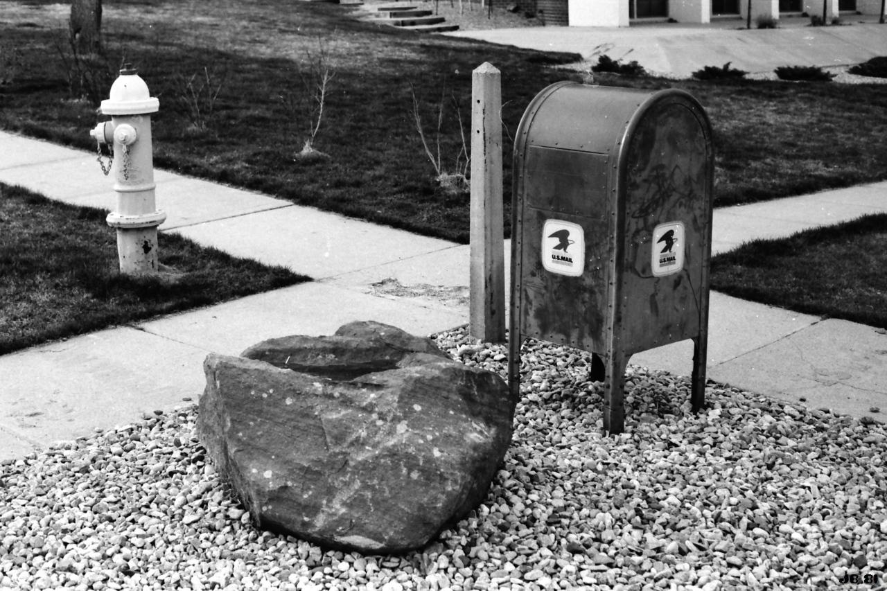 Altes Schwarzweißfoto von 1981, das zwei kreuzende Fußgängerwege aus hellen Betonplatten, einen großen Felsen im Vordergrund, dahinter rechts einen Briefkasten der US Mail und links weiter weg einen Hydranten zeigt. Rolleiflex SL35 E auf Kodak-Film.