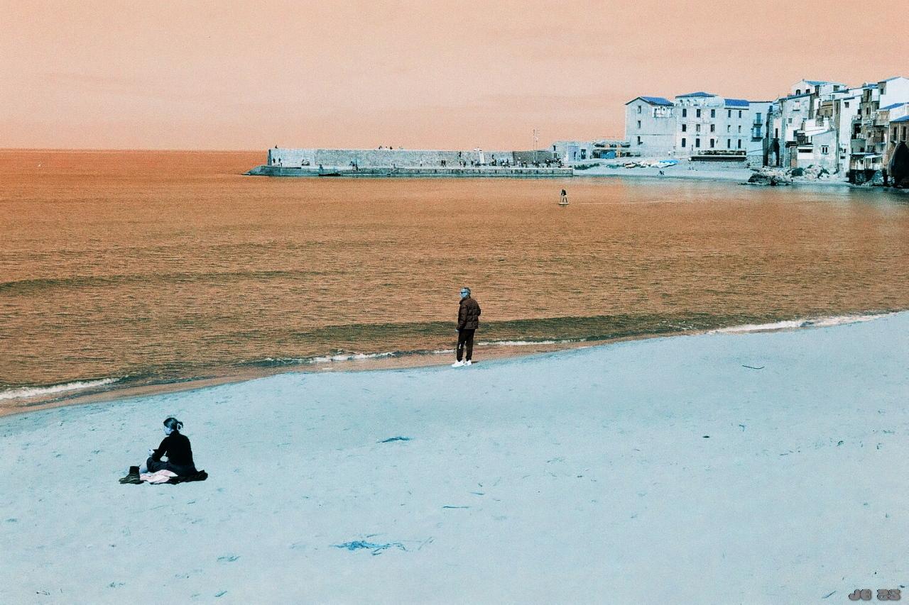 Foto in verfremdeten Farben im Braun-Türkis-Spektrum, das einen Blick auf das Mittelmeer und die Stadt Cefalù auf Sizilien zeigt. Am Strand im Vordergrund links eine meditierende Frau, in Bildmitte in Wassernähe ein Mann, im Wasser weiter entfernt ein Surfer. Leica R5 mit 50 mm Summilux auf Lomochrome Turquoise-Film.